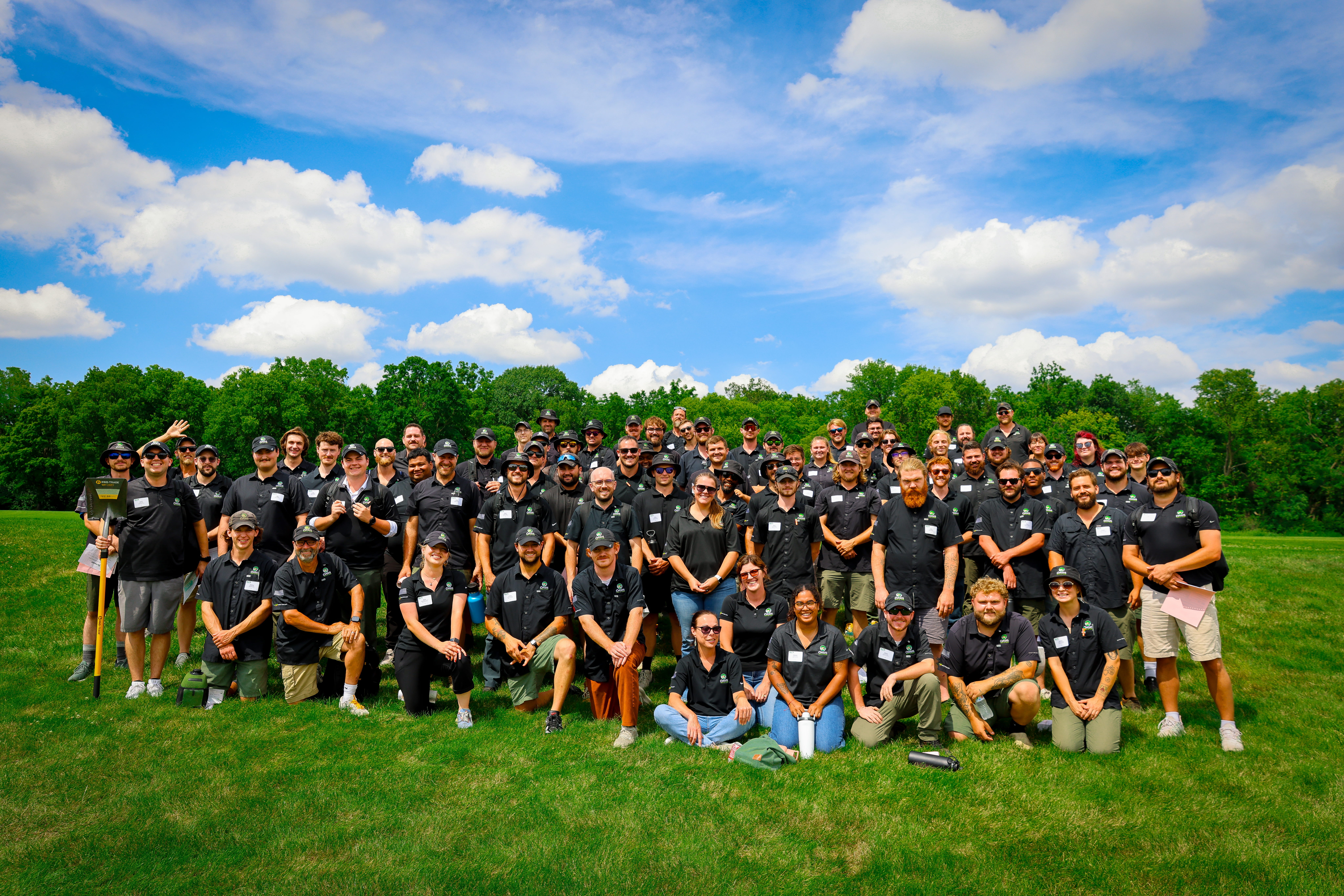 Large diverse group in matching Oasis black shirts poses on a sunny green field, smiling beneath a bright blue sky and white clouds.