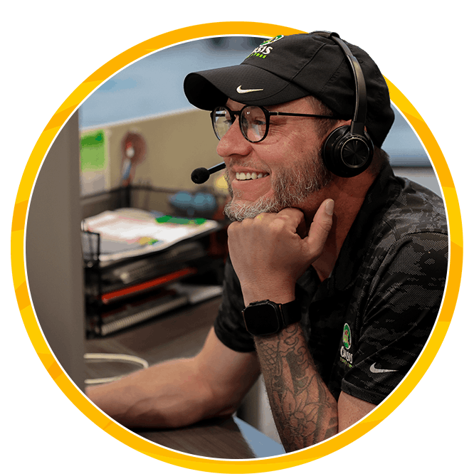 Smiling bearded man in glasses wears a headset and microphone at a desk, framed by a yellow circle; friendly, focused studio setting.