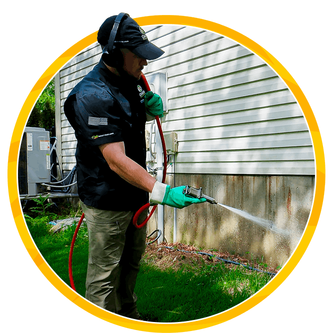Worker in protective gear spraying bifenthrin for pests outside house’s exterior siding, spraying along the foundation in a focused outdoor scene.