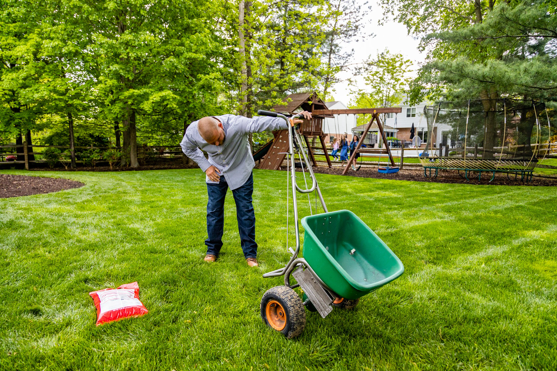 homeowner looks at fertilizer spreader while family plays in background