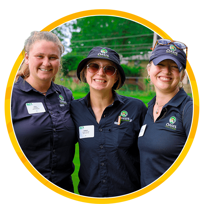 Three smiling women in matching Oasis uniforms pose outdoors inside a yellow circle, conveying teamwork, warmth, and community spirit.