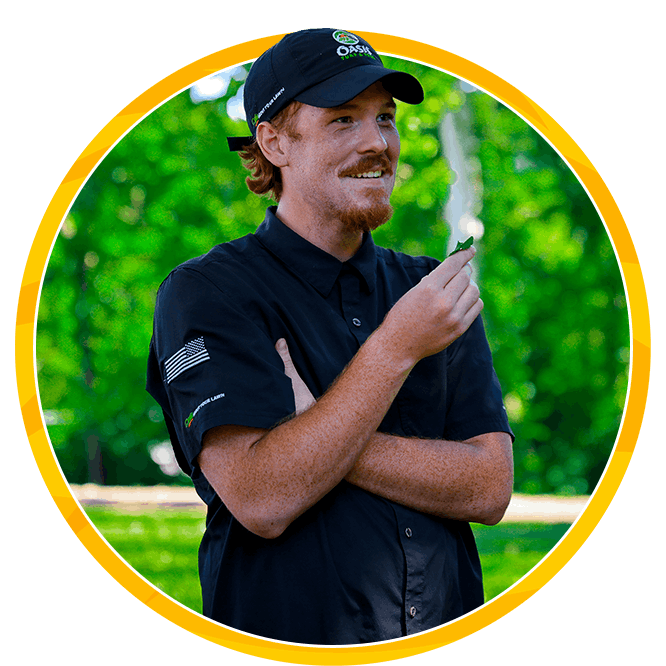 Smiling male lawn technician in a oasis black polo and cap holds a leaf outdoors, surrounded by lush green trees inside a yellow circular frame.